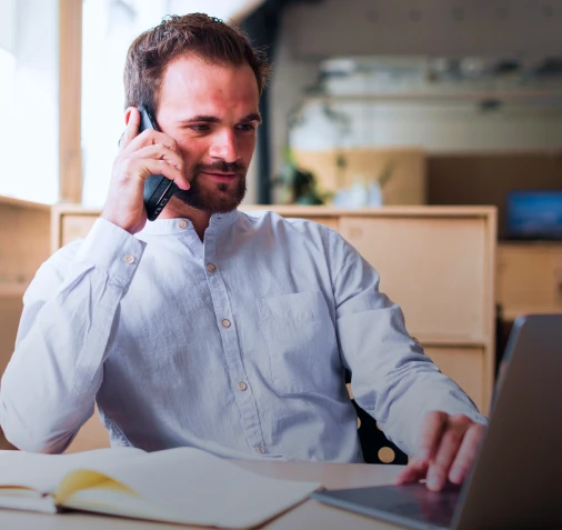 Homem vestindo social, sentando no escritório com o celular na orelha e o notebook na frente